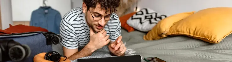 Un jeune homme aux cheveux bouclés et aux lunettes est allongé sur un lit, absorbé par le contenu d'une tablette ou d'un écran.
