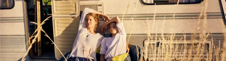 Deux personnes se détendent à l'extérieur d'une caravane par une journée ensoleillée, entourées d'herbes hautes et dorées.