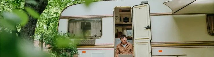 Un homme travaille sur son ordinateur portable, assis sur une terrasse en bois devant sa caravane entourée d'arbres verdoyants.