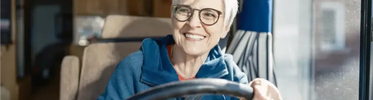 Une femme souriante, les cheveux gris, vêtue d'un pull à capuche bleu, est assise au volant de son camping-car, prête à prendre la route.