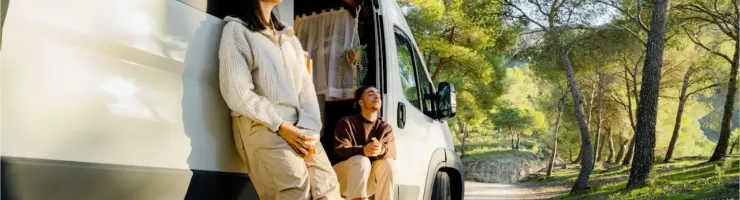 Deux jeunes se détendent à l'extérieur d'un van aménagé garé dans une forêt ensoleillée, profitant de la nature.