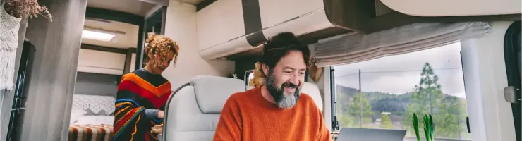 Un homme barbu utilise un ordinateur portable à l'intérieur d'un camping-car, tandis qu'une femme se tient en arrière-plan.