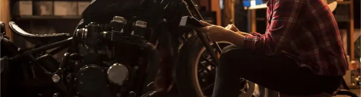 Une femme concentrée travaille sur une moto noire dans un garage, penchée sur le guidon, un outil à la main.