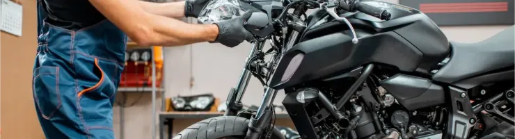 Un mécanicien en salopette bleue et gants noirs assemble le phare d'une moto noire dans un atelier.