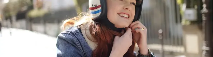 Une jeune femme rousse souriante ajuste son casque de moto vintage blanc et coloré, prête à partir.