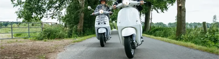 Deux personnes en casque conduisant des scooters de type Vespa sur une route de campagne bordée d'arbres.