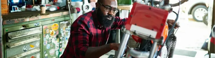 Un homme barbu avec des lunettes travaille sur une moto dans un garage encombré d'outils et de pièces.