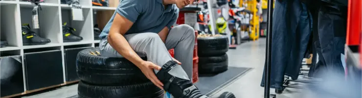 Un jeune homme essaie des bottes de moto dans un magasin, assis sur des pneus empilés.