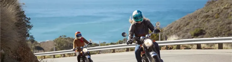 Deux femmes motardes sur leurs motos roulant sur une route côtière ensoleillée avec l'océan en fond.