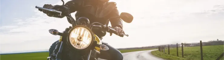 Un motard en blouson de cuir et casque roule sur une route sinueuse en campagne, au soleil couchant.