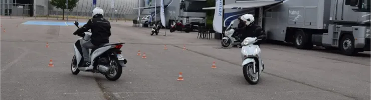 Des élèves motards en scooter pratiquent la conduite sur un parking avec des cônes, près de camions.