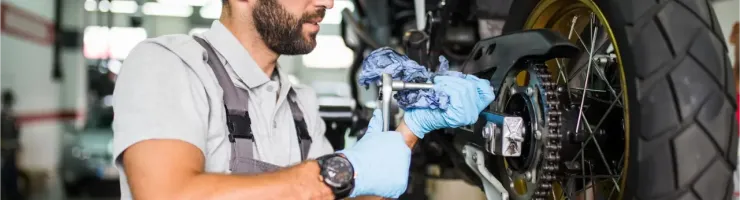Un mécanicien barbu en combinaison grise et gants bleus travaille sur la roue arrière d'une moto.