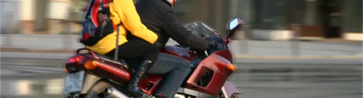 Un couple en casque de moto sur une moto rouge en ville. L'homme conduit, et la femme, avec un sac à dos, est assise derrière lui.