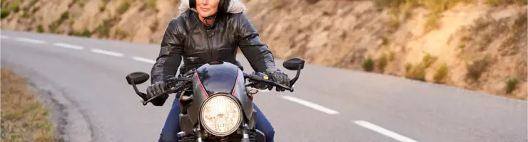 Une femme mature en blouson de cuir et casque noir conduit une moto sur une route de campagne.
