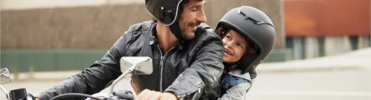 Un père souriant et son enfant en casque noir sur une moto, partageant un moment de complicité.