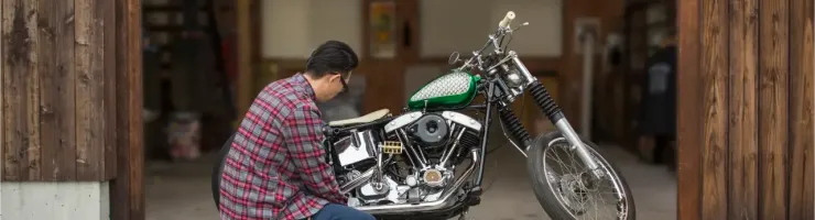 Un homme accroupi répare une moto custom dans un garage en bois ouvert avec des outils.