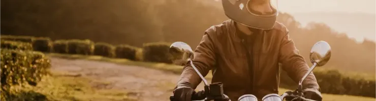 Un motard en casque rétro et blouson en cuir sur une moto vintage au coucher du soleil.