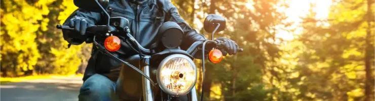 Un motard en casque et lunettes de soleil roule sur une route forestière ensoleillée.