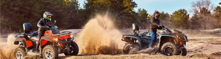 Deux personnes en quads soulèvent de la poussière en roulant sur un terrain sablonneux, avec des arbres en arrière-plan.