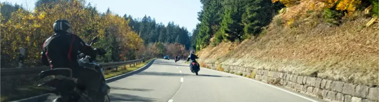 Des motards roulent sur une route sinueuse bordée d'arbres aux couleurs automnales sous un ciel clair.