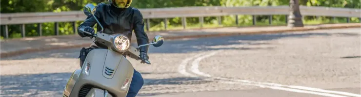 Un homme en casque et blouson de cuir conduit un scooter Vespa beige sur une route ensoleillée.