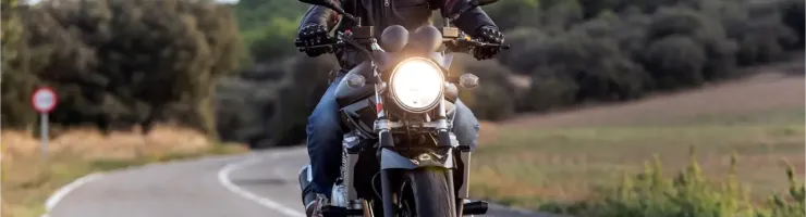 Un motard en casque noir et blouson de cuir roule sur une route de campagne sinueuse au crépuscule.