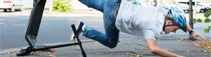 Un homme en jean et t-shirt avec un casque bleu tombe à côté de sa trottinette électrique sur un trottoir.