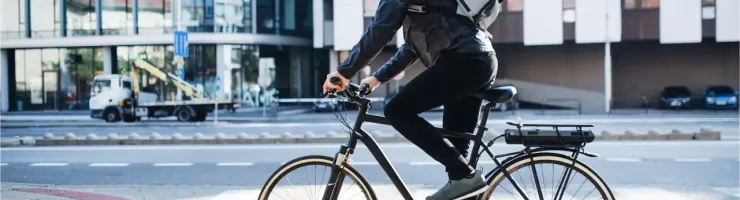 Un homme en casque et sac à dos roule sur un vélo électrique noir en ville, avec des bâtiments modernes en arrière-plan.