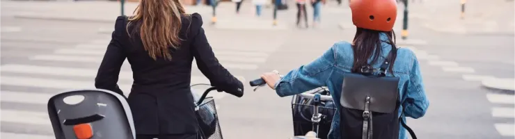 Deux femmes avec des casques sur la tête, l'une avec un siège enfant, l'autre avec un sac à dos, traversent la rue à vélo.