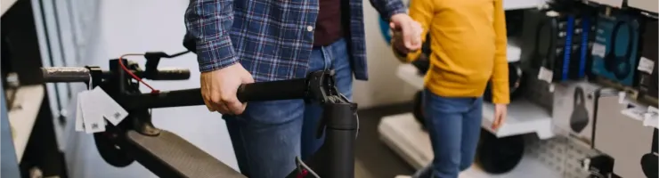 Un homme tient une trottinette électrique tandis qu'une petite fille à lunettes, souriante, lui tient la main dans un magasin.