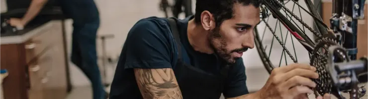 Un mécanicien barbu avec des bras tatoués répare la chaîne d'un vélo dans un atelier, un autre homme en arrière-plan.
