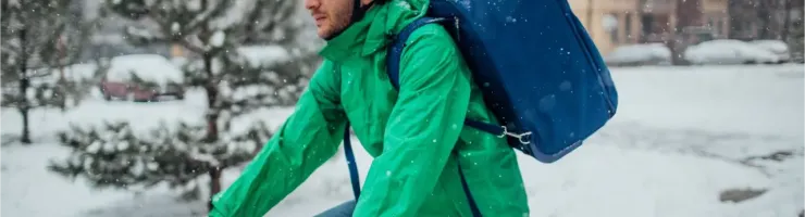 Un livreur à vélo avec un casque blanc et un grand sac bleu pédale sous la neige.