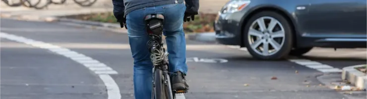 Un cycliste en doudoune et jean bleu se déplace sur une piste cyclable. On peut voir la roue arrière de son vélo et l'arrière d'une voiture.