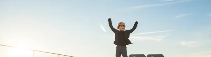 Un enfant casqué lève les bras en signe de victoire sur un hoverboard, dans un skatepark sous un ciel bleu.