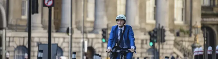 Un homme en costume et casque roule à vélo sur une rue bordée de bâtiments classiques, avec un feu rouge en arrière-plan.