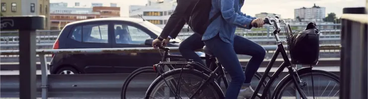 Un homme et une femme casqués roulent à vélo sur une piste cyclable en ville, avec une voiture en arrière-plan.