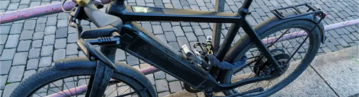 Un vélo électrique noir, avec un antivol attaché, est garé contre une barrière dans une rue pavée.