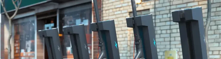 Des trottinettes électriques grises sont alignées le long d'un mur de briques, prêtes à être utilisées ou chargées.