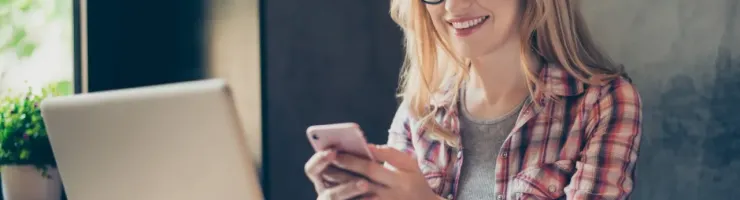 Une jeune femme souriante avec des lunettes utilise son smartphone devant un ordinateur.