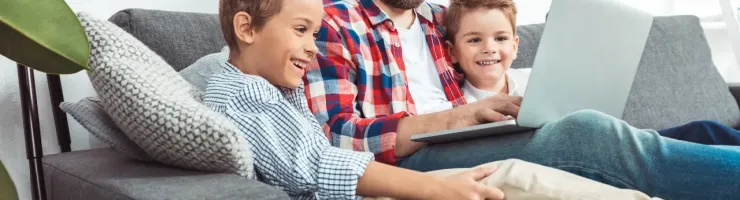 Un père et ses deux fils souriants regardent un ordinateur portable sur un canapé.