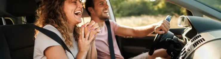 Un couple joyeux et rieur à l'intérieur d'une voiture, la femme en passagère et l'homme au volant, profitant du trajet.