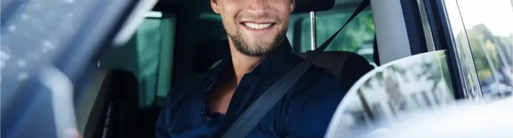 Un homme souriant et barbu conduit une voiture, regardant par la fenêtre latérale.