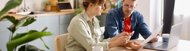 Un couple examine des papiers et un ordinateur portable, attablé dans leur cuisine. Ambiance domestique et sérieuse.