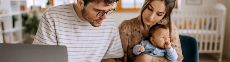 Un couple examine des documents, un ordinateur portable et une calculatrice à côté. La femme tient son nouveau-né dans ses bras.