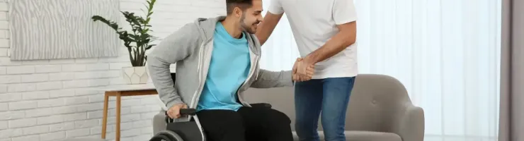 Un jeune homme aide un ami en fauteuil roulant à se lever dans un salon.
