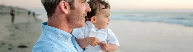 Un père de profil porte son jeune enfant dans ses bras face à la mer.