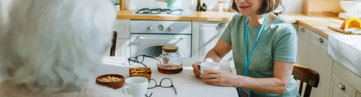 Une femme avec un badge discute chaleureusement autour d'un café avec une dame âgée dans une cuisine lumineuse.