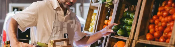 Un homme avec un tablier trie des fruits et légumes frais sur un étal en bois dans une épicerie.