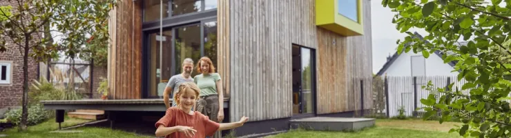Une petite fille court dans le jardin devant une maison moderne en bois, ses parents en arrière-plan.