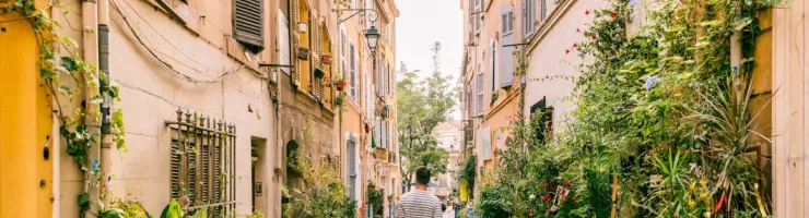 Homme marchant dans une ruelle étroite et ensoleillée de Marseille, bordée de plantes en pots et de vieux bâtiments.